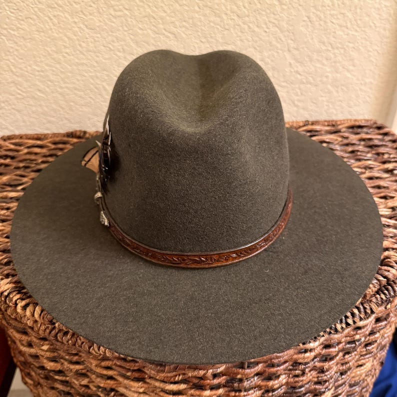 May include: A dark olive green fedora hat with a wide brim. The hat features a brown leather band with a decorative pattern and a feather accent. The hat is sitting on a woven basket.