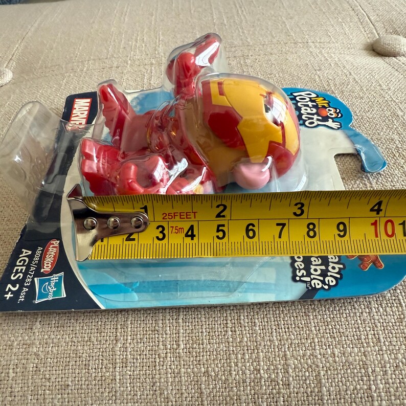 Może przedstawiać: Zabawka Marvel Mr. Potato Head w opakowaniu. Zabawka jest czerwono-ż&oacute;łta, przypominająca Iron Mana. Opakowanie zawiera logo Marvel, Playskool i markę Hasbro. Zabawka ma około 10 cm długości.