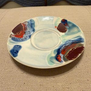 May include: A ceramic saucer with a central recessed area, painted with abstract designs in shades of blue, red, and brown against a white background. The saucer has an irregular shape and is likely handmade.