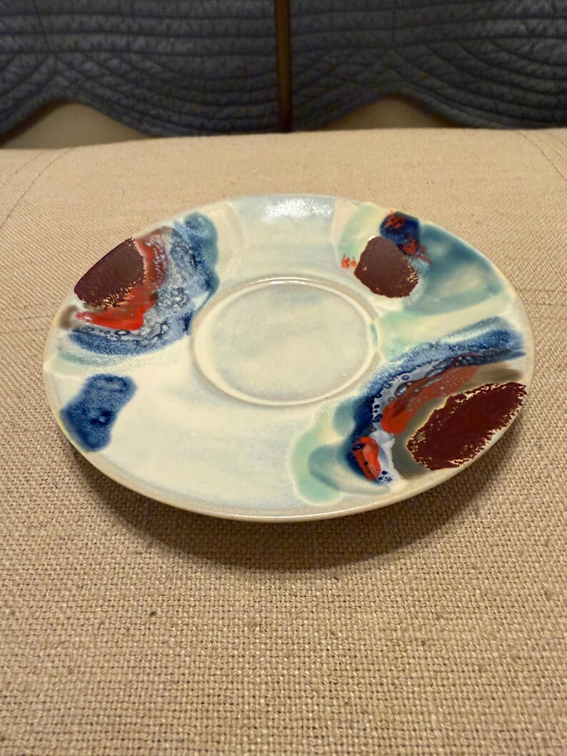 May include: A ceramic saucer with a central recessed area, painted with abstract designs in shades of blue, red, and brown against a white background. The saucer has an irregular shape and is likely handmade.