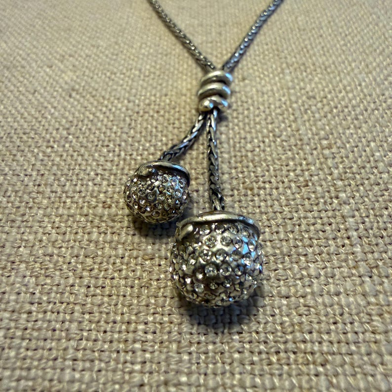 May include: A silver-toned lariat necklace featuring a braided chain that passes through a central knot, ending in two spherical pendants. Each sphere is encrusted with numerous small, clear, reflective crystals that catch the light.