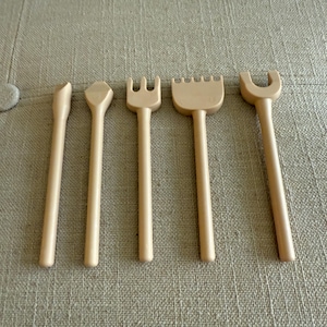 May include: Five beige sand tools, including a rake, a fork, and a U-shaped tool. The tools are made of a light-colored plastic and are arranged in a row. These tools are designed for use in sandboxes or at the beach.