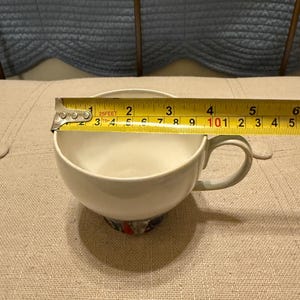 May include: A white teacup with a handle, measuring approximately 3 inches in diameter. The base of the cup features a colorful floral design. A measuring tape is placed across the cup's opening.