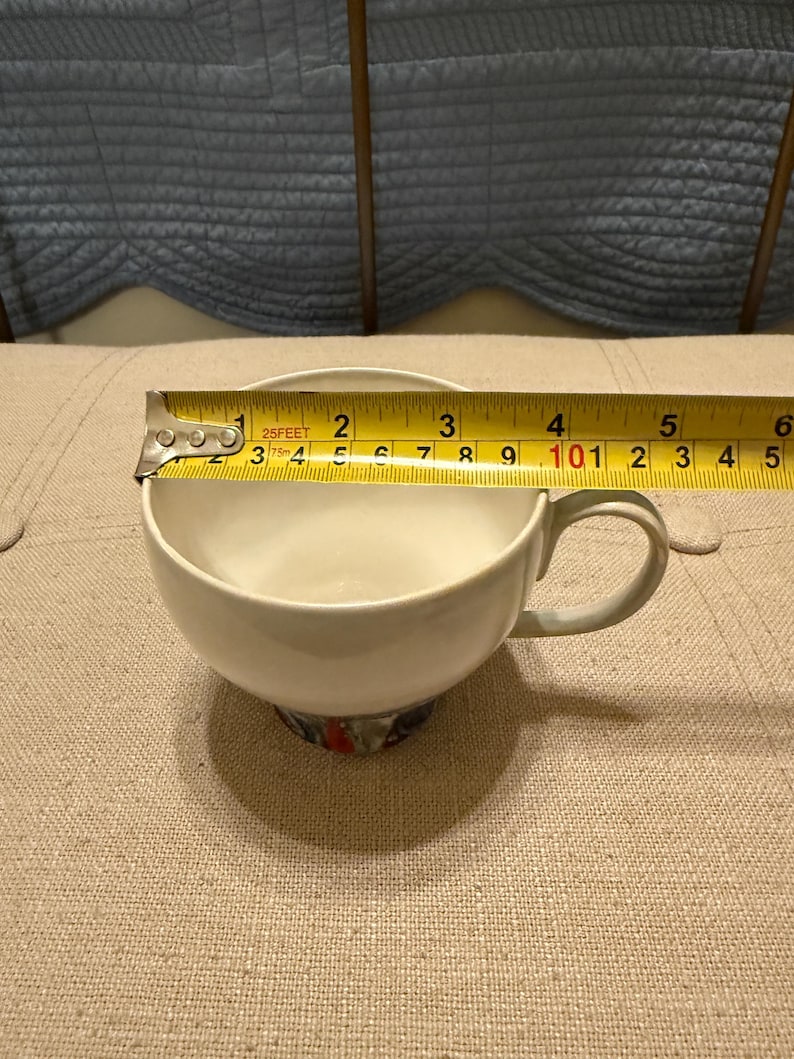 May include: A white teacup with a handle, measuring approximately 3 inches in diameter. The base of the cup features a colorful floral design. A measuring tape is placed across the cup's opening.