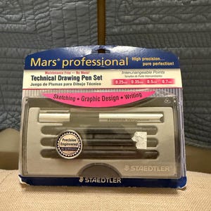 May include: A STAEDTLER Mars professional technical drawing pen set in a clear plastic package. The set includes a pen and interchangeable points in sizes 0.25, 0.35, 0.5, and 0.7 mm. The package text indicates the set is for sketching, graphic design, and writing.