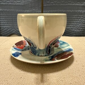 May include: A white ceramic teacup and saucer set. The cup has a large handle and is decorated with blue, red, and brown abstract designs. The saucer mirrors the cup's design. The set is on a neutral-colored surface.