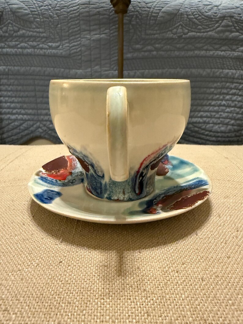 May include: A white ceramic teacup and saucer set. The cup has a large handle and is decorated with blue, red, and brown abstract designs. The saucer mirrors the cup's design. The set is on a neutral-colored surface.