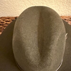 May include: A close-up of a green cowboy hat. The hat has a rounded crown and a wide brim. A thin, light-colored band encircles the base of the crown. The hat is made of a felt-like material.