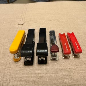 May include: A collection of six staplers in various colors, including yellow, black, and red. The staplers are arranged on a neutral-colored surface. Some staplers have the brand name "Swingline" printed on them.