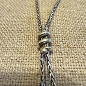 May include: A silver necklace featuring a braided chain design with a central sliding element shaped like a coiled spiral. The metal has a polished finish with darkened recessed areas that highlight the intricate texture of the woven links.