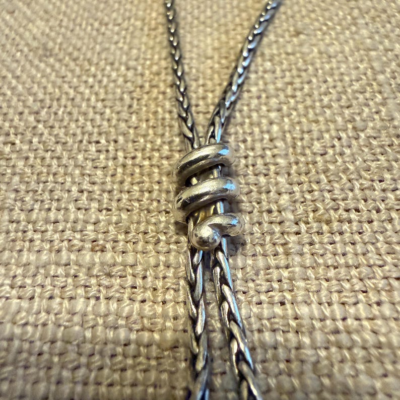 May include: A silver necklace featuring a braided chain design with a central sliding element shaped like a coiled spiral. The metal has a polished finish with darkened recessed areas that highlight the intricate texture of the woven links.