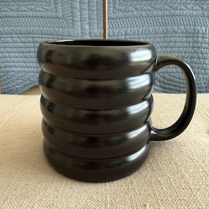 May include: A black ceramic mug with a unique, ridged design. The mug has a large handle and a glossy finish. The mug is sitting on a beige surface, with a blue quilted background.