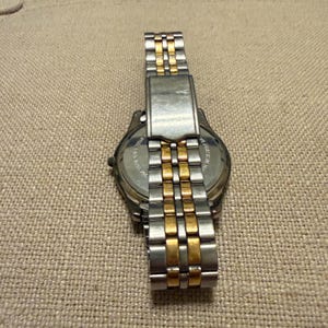 以下が含まれることがあります： シルバーとゴールドのセグメントが交互に並ぶツートンカラーの金属製リンクバンドを備えた腕時計。時計の裏蓋は円形で金属製であり、目に見えるテキストの刻印と折りたたみ式のクラスプ機構が含まれています。