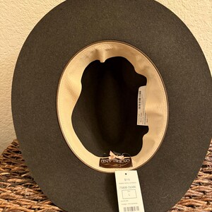 May include: A dark olive green hat with a wide brim, viewed from the inside. The hat has a light tan interior lining and a tag with the brand name "OVERLAND" and product information. The hat is a size L.