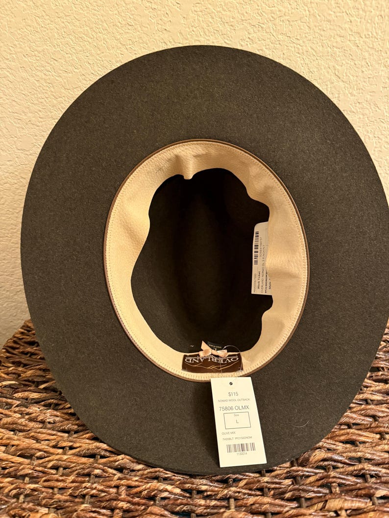 May include: A dark olive green hat with a wide brim, viewed from the inside. The hat has a light tan interior lining and a tag with the brand name "OVERLAND" and product information. The hat is a size L.