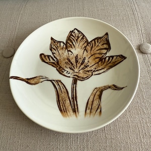 May include: A white ceramic plate featuring a brown tulip design. The tulip is centrally positioned, with detailed petals and leaves. The plate has a simple, elegant design, suitable for serving food or as a decorative piece.