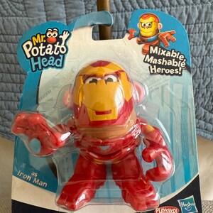 Może przedstawiać: Zabawka Mr. Potato Head jako Iron Man w opakowaniu. Zabawka jest czerwona i złota, z designem hełmu Iron Mana. Opakowanie zawiera tekst "Mixable, Mashable Heroes!"