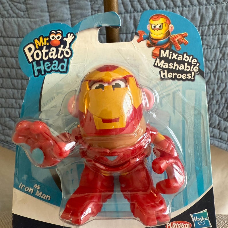 Może przedstawiać: Zabawka Mr. Potato Head jako Iron Man w opakowaniu. Zabawka jest czerwona i złota, z designem hełmu Iron Mana. Opakowanie zawiera tekst "Mixable, Mashable Heroes!"