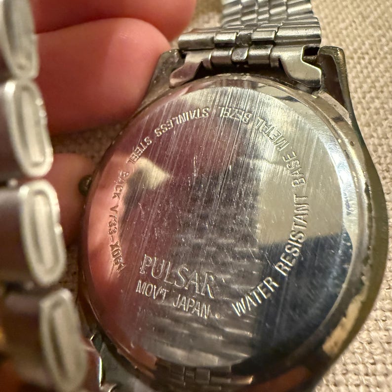 以下が含まれることがあります： 金属製のリンクバンドが付いたシルバーカラーのPulsar腕時計。時計ケースの裏蓋はステンレススチール製で、Pulsar、Movt Japan、Water Resistant、Base Metal Bezel、モデル番号V733-X061という刻印が施されています。