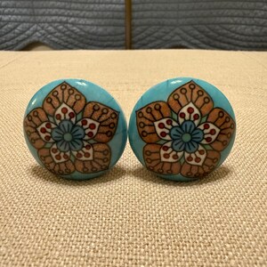 May include: Two turquoise ceramic cabinet knobs with a floral design. The flower has a brown outline with white and red accents, and a blue center. The knobs are round and appear to be new.