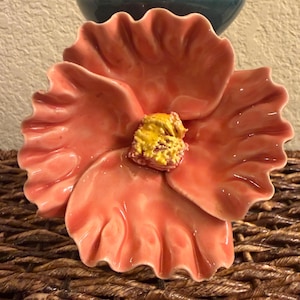 May include: A ceramic flower-shaped dish with a glossy finish. The petals are a coral color, and the center has a textured yellow and orange design. The dish is placed on a woven surface, with a glimpse of a blue vase in the background.