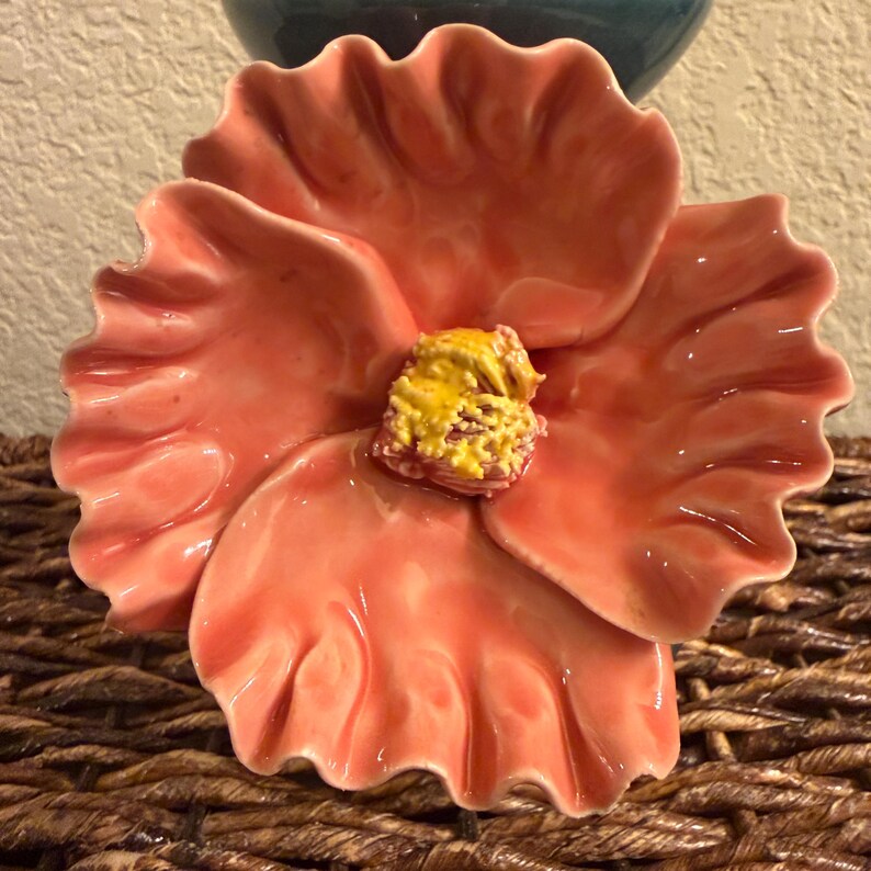 May include: A ceramic flower-shaped dish with a glossy finish. The petals are a coral color, and the center has a textured yellow and orange design. The dish is placed on a woven surface, with a glimpse of a blue vase in the background.