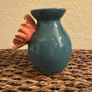 May include: Teal ceramic vase with a pink flower accent. The vase has a rounded body and a flared rim. The flower is attached to the side of the vase and has multiple petals. The vase is sitting on a woven surface.