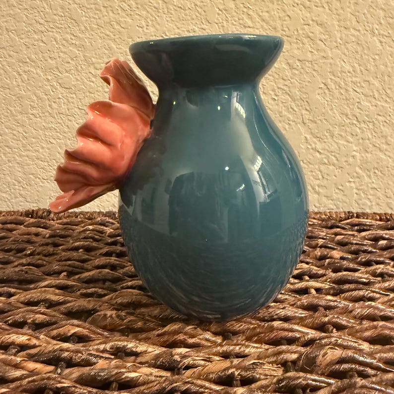 May include: Teal ceramic vase with a pink flower accent. The vase has a rounded body and a flared rim. The flower is attached to the side of the vase and has multiple petals. The vase is sitting on a woven surface.