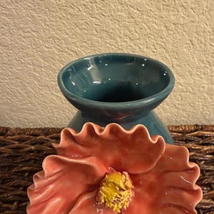 May include: A teal ceramic bowl with a flared base and a coral-colored, flower-shaped dish. The flower has a yellow center and ruffled edges. The items are on a woven surface.