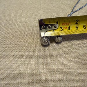 May include: A silver-toned necklace featuring two spherical pendants encrusted with small clear stones. A yellow measuring tape is positioned over the pendants, showing the larger sphere measures approximately 0.5 inches in diameter.