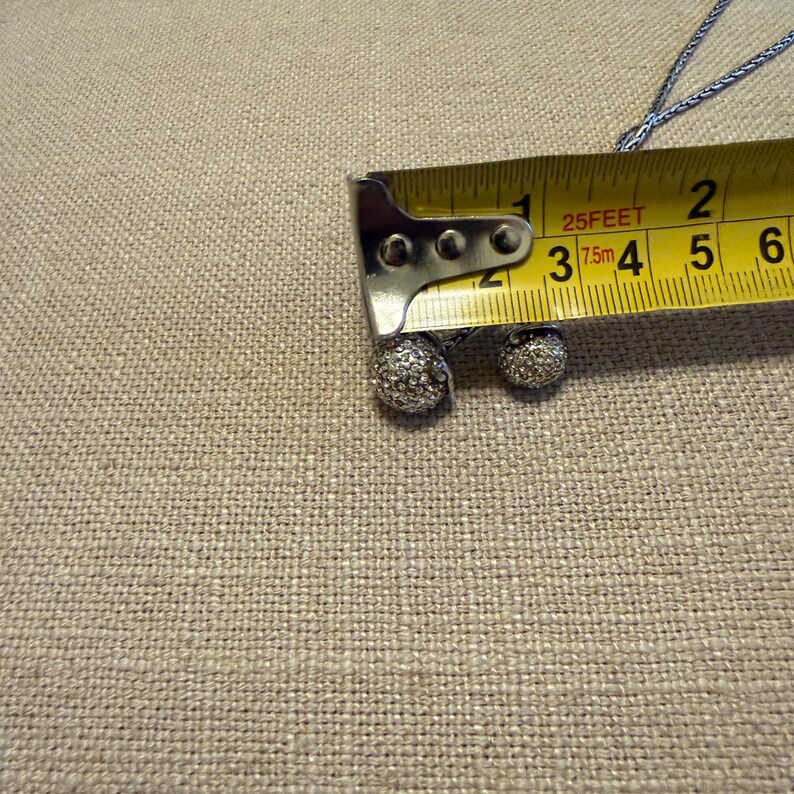 May include: A silver-toned necklace featuring two spherical pendants encrusted with small clear stones. A yellow measuring tape is positioned over the pendants, showing the larger sphere measures approximately 0.5 inches in diameter.