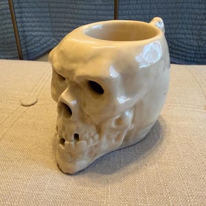 May include: A cream-colored ceramic skull-shaped mug. The mug has a large opening at the top and a small handle. The skull features detailed facial features, including eye sockets, a nose cavity, and teeth. The mug is a unique and decorative item.