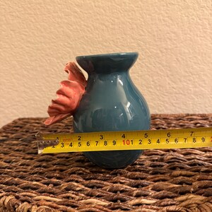 May include: A teal ceramic vase with a pink floral accent. The vase is approximately 4 inches in diameter and is sitting on a woven surface. The vase has a glossy finish.