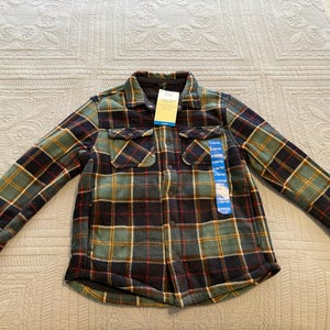May include: A plaid flannel shirt in shades of green, navy, red, and yellow. The shirt has two chest pockets and a button-down front. A tag is attached to the shirt.