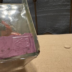 以下が含まれることがあります： 紫と白のパターンが施された透明なプラスチック製の箱。箱は部分的に開いており、中に小さな茶色のフィギュアが入っている様子が見えます。