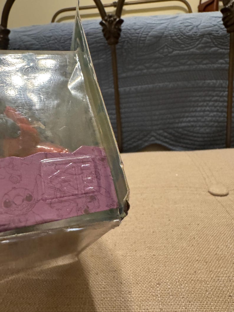 以下が含まれることがあります： 紫と白のパターンが施された透明なプラスチック製の箱。箱は部分的に開いており、中に小さな茶色のフィギュアが入っている様子が見えます。