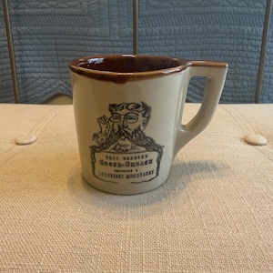 May include: A beige ceramic mug with a brown rim and handle. The mug features a black and white illustration of a mustached man with glasses and the text "PROX. BARBER GOOSE-GREASE PRODUCES A LUXURIANT MOUSTACHE."