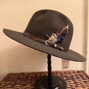 May include: A dark green fedora-style hat with a wide brim. The hat features a brown leather band with a decorative buckle and is adorned with feathers in shades of brown and blue. The hat is displayed on a black stand.