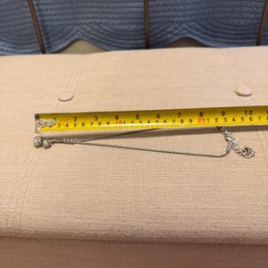 May include: A silver-toned chain necklace featuring two small spherical charms at one end and a lobster claw clasp at the other. A yellow measuring tape is positioned above the necklace, indicating a length of approximately 10 inches.