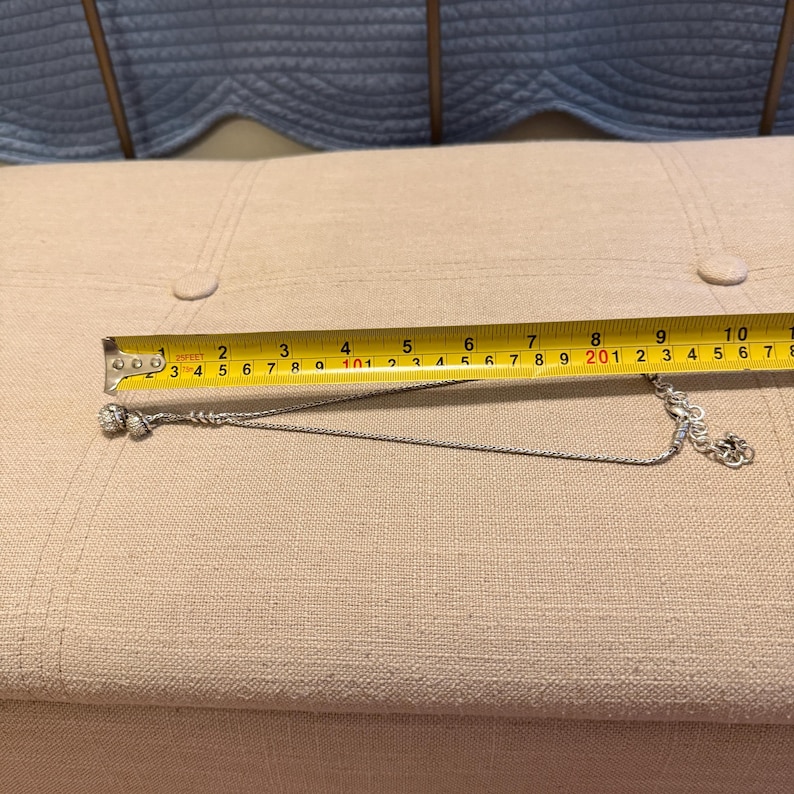May include: A silver-toned chain necklace featuring two small spherical charms at one end and a lobster claw clasp at the other. A yellow measuring tape is positioned above the necklace, indicating a length of approximately 10 inches.