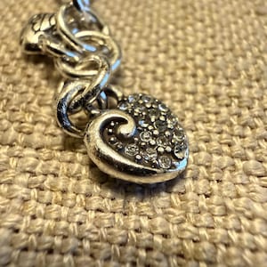 May include: A silver-toned charm bracelet featuring a heart-shaped pendant with a swirling design and embedded clear crystals. The charm is attached to a thick, rounded silver-toned chain link, showcasing a metallic finish and reflective surfaces.