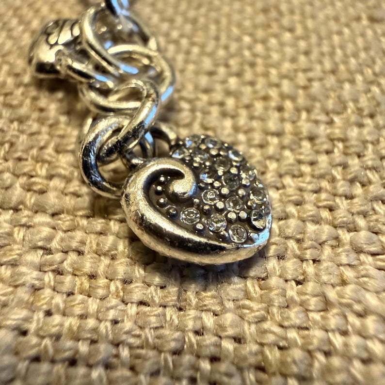 May include: A silver-toned charm bracelet featuring a heart-shaped pendant with a swirling design and embedded clear crystals. The charm is attached to a thick, rounded silver-toned chain link, showcasing a metallic finish and reflective surfaces.