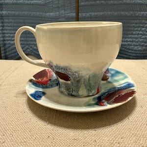 May include: A ceramic teacup and saucer set. The cup is off-white with blue and red abstract paint strokes. The saucer has matching blue and red abstract paint strokes. The set is on a beige surface.