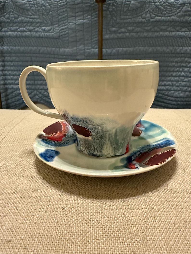 May include: A ceramic teacup and saucer set. The cup is off-white with blue and red abstract paint strokes. The saucer has matching blue and red abstract paint strokes. The set is on a beige surface.