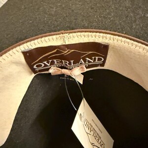 May include: A dark gray hat with a brown leather band and a cream-colored interior. The hat features a tag that reads "OVERLAND ESTABLISHED 1973". The hat is made of wool.