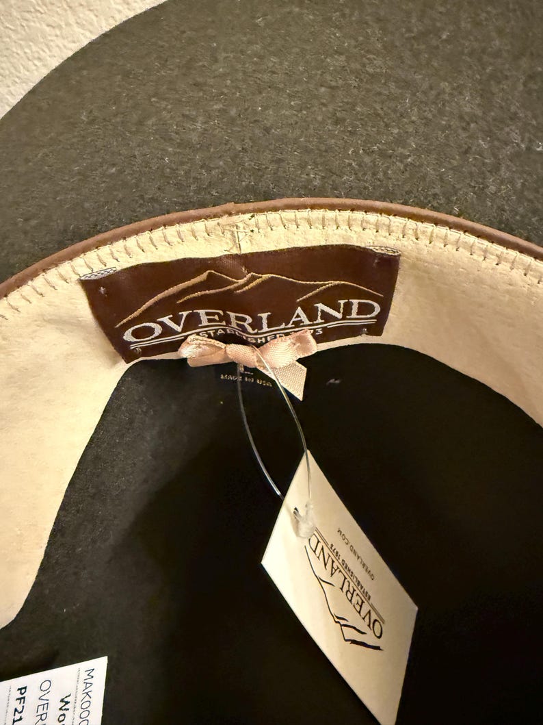 May include: A dark gray hat with a brown leather band and a cream-colored interior. The hat features a tag that reads "OVERLAND ESTABLISHED 1973". The hat is made of wool.