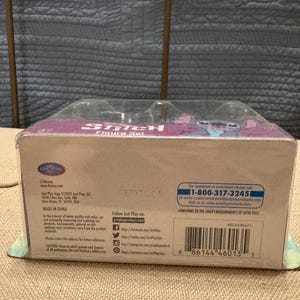 以下が含まれることがあります： スティッチのフィギュアセットが入った青と紫の箱。箱の前面にはディズニーのロゴと「スティッチフィギュアセット」の文字が書かれている。箱の裏面には、製造元、ウェブサイト、安全に関する情報など、製品に関する情報が記載されている。バーコードは箱の裏面に表示されている。