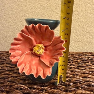 May include: A teal ceramic vase with a large, coral-colored flower attached. The flower has a yellow center. The vase is approximately 5 inches tall, as measured by a yellow tape measure.