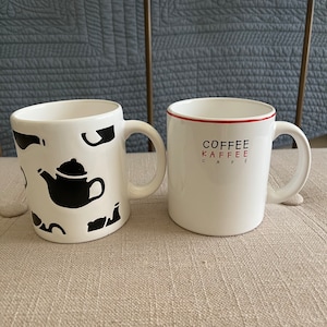 May include: Two white ceramic mugs. One mug has a black silhouette of a teapot and other teapots. The other mug has the words "COFFEE KAFFEE CAFE" in red and black.