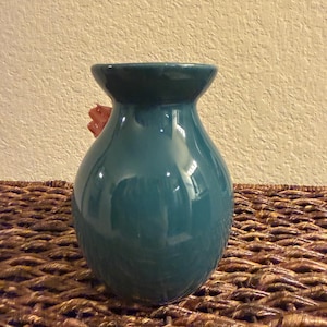 May include: A teal ceramic vase with a glossy finish. The vase has a rounded body and a flared opening. A red flower is partially visible inside the vase. The vase is sitting on a woven brown surface.